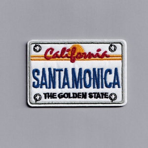 Peut inclure: Un patch blanc avec un design de plaque d'immatriculation californienne brodé. La plaque indique "California" avec un soleil jaune et "Santa Monica" en bleu. Le bas de la plaque indique "The Golden State" en noir.