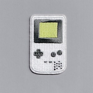 Op de afbeelding: Witte en grijze geborduurde patch van een draagbare videogameconsole. De console heeft een zwart-wit scherm met een groen gepixelde display. De console heeft een directionele pad, twee knoppen en een startknop.