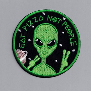 Op de afbeelding: Groen buitenaards wezen met een vredesteken en de tekst "Eat Pizza Not People" op een zwarte achtergrond met witte sterren.