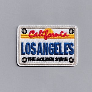 以下が含まれることがあります： 「カリフォルニア」の文字と黄色と赤の夕焼けのグラフィックが施された白と青の刺繍パッチ。「ロサンゼルス」の文字は青、「ザ・ゴールデン・ステート」の文字は黒です。