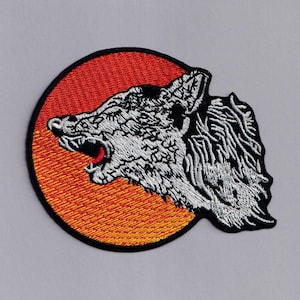 May include: An embroidered patch depicting a white wolf howling at a red and orange sun. The wolf is detailed with white stitching and the sun has a textured orange and yellow design.