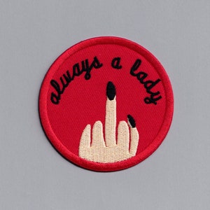 May include: Red round embroidered patch with a white hand giving the middle finger and the text "always a lady" in black.