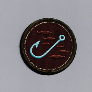 Può includere: Una toppa ricamata rotonda di colore marrone e verde con un amo da pesca blu e uno scuola di pesci marroni. La toppa è circolare e ha un bordo cucito.