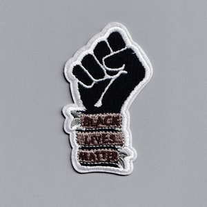 Peut inclure: Patch brodé "Black Lives Matter" avec un poing levé en noir et un contour blanc. Les mots "Black Lives Matter" sont enroulés autour du poignet du poing en fil brun.