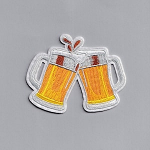 May include: Two white ceramic beer mugs with yellow beer and brown foam, clinking together. The mugs have a white outline and are embroidered.