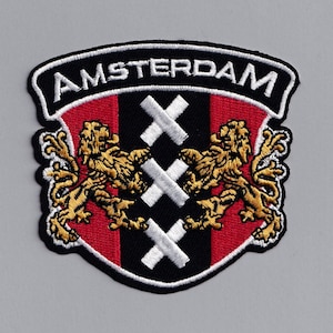 Puede incluir: Un parche bordado en blanco y negro con la palabra "AMSTERDAM" en un arco blanco sobre un escudo rojo, negro y blanco con dos leones dorados y cruces blancas.