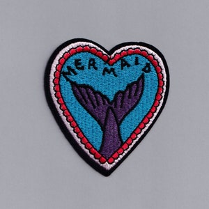 May include: A heart-shaped embroidered patch with a blue background and a purple mermaid tail. The words "MERMAID" are embroidered in white around the edge of the heart.