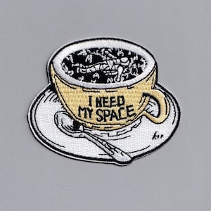 May include: An embroidered iron-on patch featuring a yellow coffee cup with a black outline and the text "I NEED MY SPACE". The cup is filled with black coffee and a small astronaut is floating in the coffee.