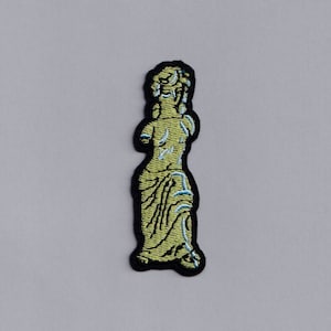 Könnte beinhalten: Ein gestickter Aufnäher, der eine Statue einer Frau darstellt, in Gelb mit blauer Umrandung. Der Aufnäher befindet sich auf einem weißen Hintergrund.