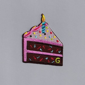 May include: A brown and pink embroidered patch of a slice of chocolate cake with pink frosting and sprinkles. The cake has a lit yellow candle on top and the letter "G" embroidered on the side.