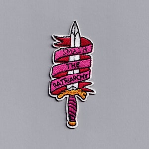 Op de afbeelding: Roze en rode geborduurde patch met een zwaard en een lint met de tekst "Smash the Patriarchy".