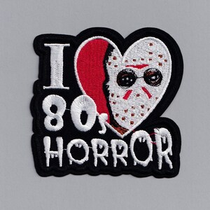 May include: Black embroidered patch with a white heart featuring a masked figure with sunglasses. The text "I 80's HORROR" is embroidered in white.
