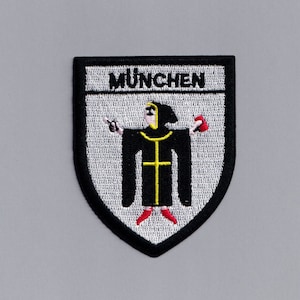 May include: A white and black embroidered patch with the word "MÜNCHEN" at the top and a black figure in a white robe with a yellow cross and red shoes holding a red object in one hand and a white object in the other hand.