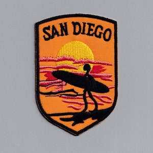 May include: An orange and black embroidered patch featuring a sunset scene and a silhouette of a surfer. The text "SAN DIEGO" is embroidered in black on the patch.