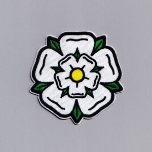 Może przedstawiać: Biała haftowana naszywka z białą różą z żółtym środkiem i zielonymi liśćmi. Róża jest obwiedziona czarną nicią.