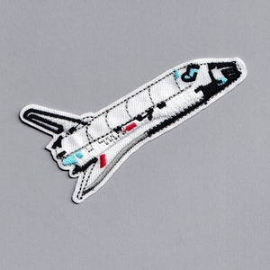以下が含まれることがあります： 黒と赤のディテールが付いた白い刺繍パッチのスペースシャトル。シャトルは鼻を上に向けて飛んでいます。
