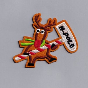 Pode incluir: Patch bordado com uma rena de desenho animado com chifres castanhos, um cachecol verde e um nariz vermelho. A rena segura uma bengala de doces e uma placa com a inscrição "N. POLE". O patch tem uma borda marrom sobre um fundo cinza.
