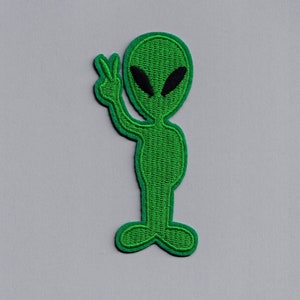 Peut inclure: Un extraterrestre vert avec des yeux noirs faisant le signe de la paix. L'extraterrestre est brodé sur un fond blanc.