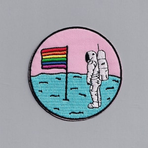 Pode incluir: Um remendo redondo bordado com um fundo rosa, uma bandeira arco-íris e um astronauta em pé sobre uma superfície azul.