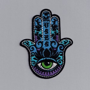 May include: An embroidered hamsa hand in blue and purple with a green eye in the centre. The hamsa hand is a symbol of protection and good luck.