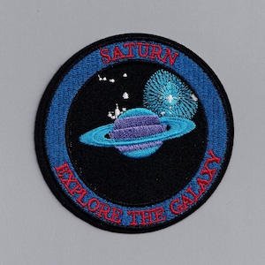 May include: A blue and black embroidered patch with the text "Saturn Explore the Galaxy" in red. The patch features a stylized image of the planet Saturn with its rings.