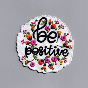 May include: White embroidered patch with a floral border and the text "be positive" in black.