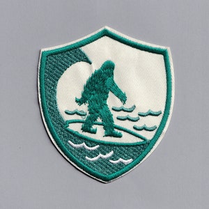 Op de afbeelding: Groen geborduurd patch in de vorm van een schild met een witte achtergrond. De patch toont een groen silhouet van een Bigfoot die op een golf surft.