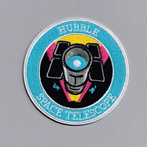 May include: A blue and white embroidered patch with the text "Hubble Space Telescope" and a stylised image of the Hubble Telescope in the centre. The telescope is surrounded by a yellow, pink and black design.