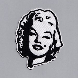 May include: Black and white embroidered patch of a woman's face with a slight smile. The woman has short hair and is wearing a simple outfit.