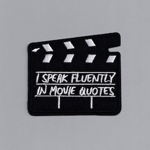 Può includere: Toppa ricamata in bianco e nero con un design di ciak cinematografico. Il testo recita "Parlo fluentemente in citazioni di film".