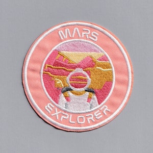 May include: Pink embroidered patch with a white outline. The patch features a stylised image of a Martian landscape with a white astronaut helmet in the foreground. The text "MARS EXPLORER" is written in a white circular border around the image.