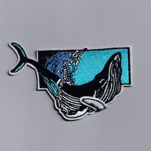 以下が含まれることがあります： 水から飛び出す白黒のクジラの刺繍パッチ。水は青とターコイズの色合いで描かれています。