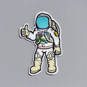 Peut inclure: Patch brodé blanc d'un astronaute faisant un pouce en l'air, portant une combinaison spatiale blanche avec une visière bleue et une bonbonne d'oxygène jaune et verte.