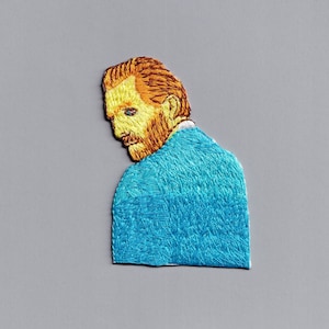 May include: Embroidered patch of a man with a red beard and blue shirt. The man is looking to the side and has a serious expression.