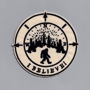 May include: A black and white embroidered patch featuring a compass design and a silhouette of Bigfoot walking through a forest. The text "I BELIEVE!" is written around the edge of the patch.