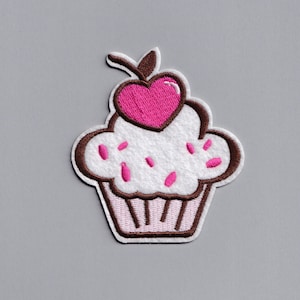 Può includere: Un cupcake bianco con glassa rosa e codette, sormontato da una ciliegia rosa a forma di cuore. Il cupcake è bordato di marrone.