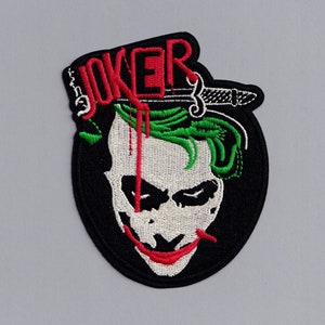 May include: Black and white embroidered patch featuring a green and white Joker face with a red smile and a red and white knife sticking out of the top of his head. The word "JOKER" is embroidered in red above the face.