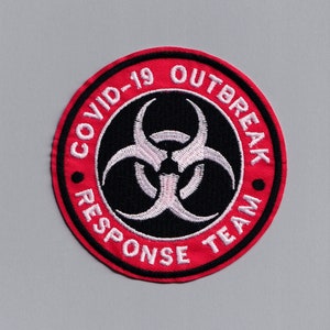 Puede incluir: Un parche circular rojo y negro con el texto "COVID-19 OUTBREAK RESPONSE TEAM" y un símbolo de riesgo biológico blanco en el centro.