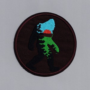 May include: A round, brown embroidered patch featuring a silhouette of Bigfoot walking. The Bigfoot is filled with green and blue colours, and a red sun is visible in the upper part of the silhouette.