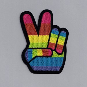 May include: A rainbow peace sign patch with a black outline. The patch is made of embroidered fabric and features a peace sign with a rainbow pattern.