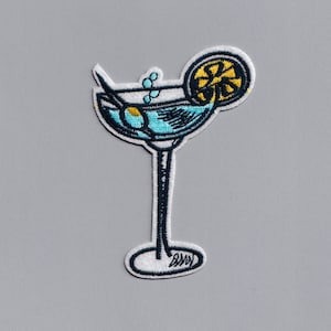 May include: An embroidered patch of a blue martini glass with a green olive and a yellow lemon wedge. The glass has a black stem and a white base with the word "BRAVO" written on it.