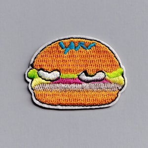 May include: A cartoon embroidered patch of a yellow hamburger with green lettuce, pink meat, and white sesame seeds on top.