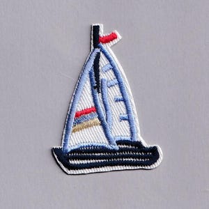 Könnte beinhalten: Ein gestickter Aufbügler in Form eines Segelboots. Das Boot hat einen dunkelmarineblauen Rumpf, weiße Segel mit hellblauen Umrandungen und horizontale Akzentstreifen in Rot, Blau und Beige am unteren Segelbereich. Das Design wird durch eine kleine rote Flagge an der Mastspitze abgerundet.