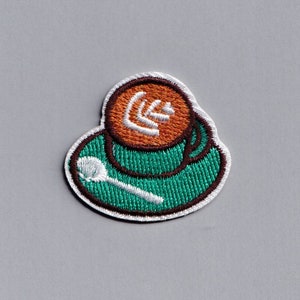 May include: An embroidered patch of a green coffee cup with a brown rim and orange coffee with a white swirl design. A white spoon is on a green saucer.