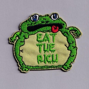 May include: An embroidered patch featuring a green cartoon frog with a pale yellow belly. The frog has blue eyes, a wide black mouth, and a small red tongue sticking out. The text EAT THE RICH is stitched in green on the frog's belly.