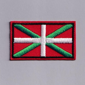 May include: A rectangular embroidered patch featuring the Ikurrina flag design. The patch has a red background with a white horizontal cross and a green diagonal cross layered over it, all enclosed within a thick black stitched border.
