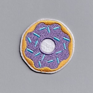 Könnte beinhalten: Lilafarbener Donut mit blauen Streuseln und einem weißen Zentrum. Der Donut ist auf einem weißen Stoff gestickt.