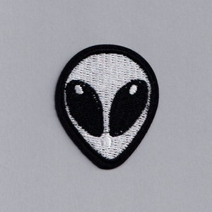 Op de afbeelding: Een geborduurde patch met een wit alien gezicht met zwarte ogen. De patch is op een grijze achtergrond.