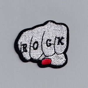 Może przedstawiać: Biała haftowana łatka przedstawiająca pięść z napisem "ROCK" na kostkach. Pięść jest zaciśnięta, a palec wskazujący ma czerwony lakier do paznokci.