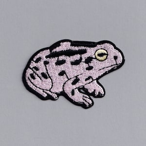 Może przedstawiać: Naszywka z haftowanej różowej żaby z czarnymi konturami i żółtym okiem.